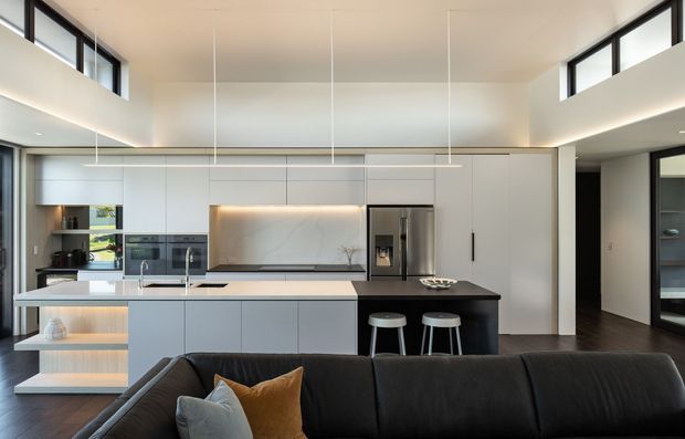 A bright, open-plan contemporary kitchen viewed from the living area. The kitchen features a large pale island with integrated sink and seating, dark contrasting cabinetry and benchtop, tall white cabinetry, stainless-steel appliances, open shelving, and concealed warm under-cabinet lighting. High clerestory windows and a raised ceiling create an airy, spacious feel, while a dark sectional sofa occupies the foreground.