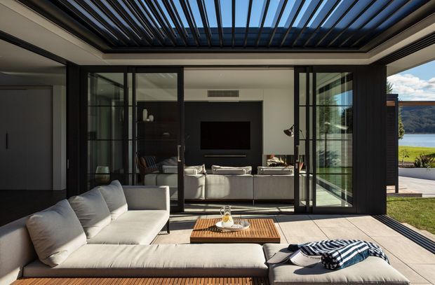 A sophisticated indoor-outdoor living area with a large upholstered sectional and chaise on a tiled terrace, arranged around a timber slatted coffee table. Fully glazed black-framed folding or sliding doors open toward an interior living room with a television and sofa. Overhead, a striking louvred roof provides shade while retaining views of the bright waterfront landscape beyond.
