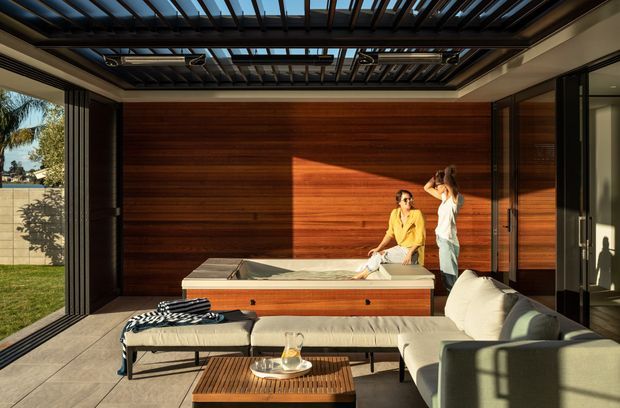 A warm, contemporary indoor-outdoor living space with a large built-in spa or plunge pool set against a rich horizontal timber feature wall. A louvred pergola-style ceiling brings in sunlight, while full-height sliding glass doors open the room to a lawn and landscaped exterior. The room includes a pale sectional sofa, ottoman seating, and a timber coffee table.