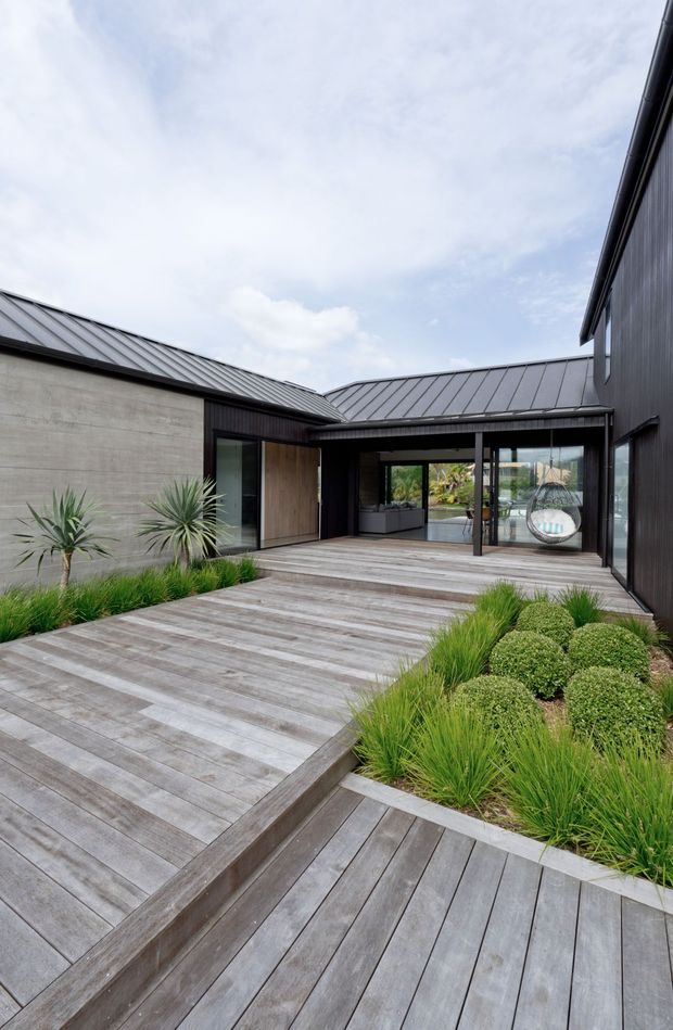 Contemporary low-rise courtyard exterior with dark vertical cladding, grey metal pitched roofs, a raw concrete wall, extensive timber decking and stepped boardwalks, landscaped grasses and sculptural planting, and large glazed openings connecting to an outdoor seating area.