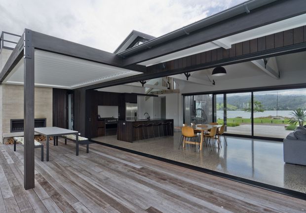 A contemporary open-plan indoor-outdoor living area with a large timber deck, covered outdoor dining zone, dark-framed sliding glass doors, and a bright interior containing a kitchen island and dining table. Exposed structural beams and a pitched roof form are visible, with a landscaped waterfront or lake view beyond the glazing.