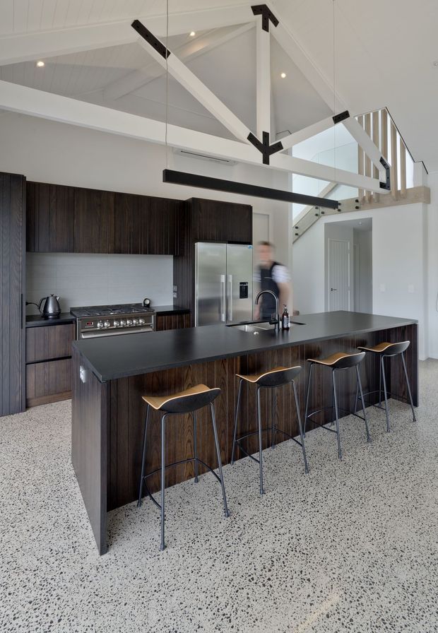 A bright, contemporary kitchen with a large dark island and integrated seating for four. The room features dark vertical timber cabinetry, a pale tiled splashback, stainless-steel appliances, a polished terrazzo floor, and a dramatic pitched ceiling with exposed white structural beams accented by black metal connectors. A stair and upper-level balustrade are visible beyond the kitchen.