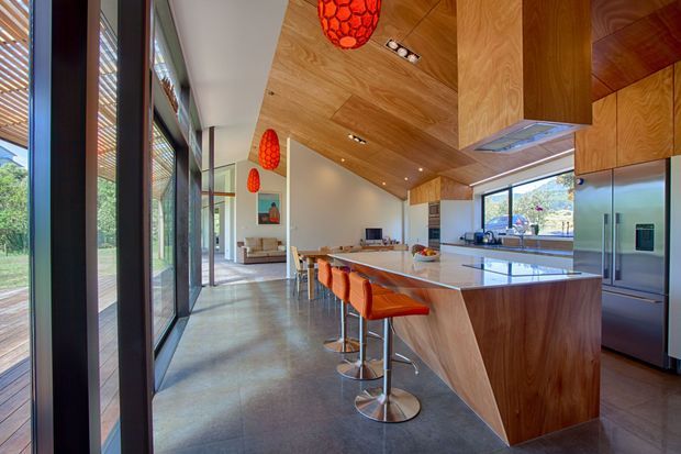 A spacious open-plan kitchen and living area with a large timber-clad island and integrated cabinetry, polished concrete flooring, full-height dark-framed glazing, and a dramatic timber ceiling. Orange upholstered bar stools and red pendant lights provide vivid accents, while the glazing opens toward a leafy outdoor setting.