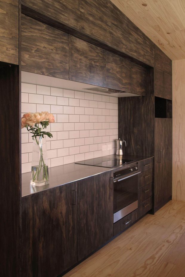 Compact galley-style kitchen with dark wood-grain full-height cabinetry, integrated rangehood, black induction cooktop and built-in oven. A pale pink-white subway-tile splashback provides contrast, while the surrounding walls and ceiling are finished in light natural timber. The space has a warm, refined, contemporary feel.