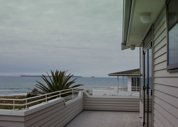 Exterior coastal terrace overlooking the ocean, with pale horizontal weatherboard cladding, white railings, a broad overhanging eave, glazed doors, and raised planter beds containing palm-like planting. The scene has an overcast, muted seaside atmosphere.