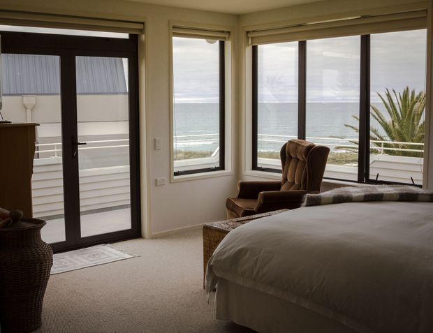 A warm, coastal bedroom with a large bed, upholstered vintage-style armchair, woven furniture, and carpeted floor. Corner windows and a glazed door provide expansive ocean views, with a small balcony or exterior landing visible beyond the glazing. Neutral beige and brown tones create a calm, inviting atmosphere.