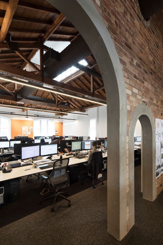 Open-plan commercial office interior in a converted industrial-style space, featuring rows of workstations, computer monitors, task chairs, exposed timber roof framing, exposed services, skylights, white walls with an orange feature wall, and a prominent red brick partition with large arched openings finished in grey plaster.