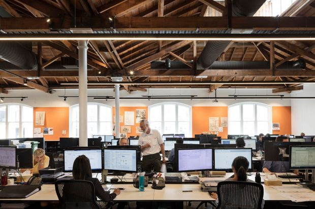 Open-plan commercial office interior with rows of workstations, computer monitors, office chairs, and employees. The space features exposed timber roof trusses and beams, exposed services and ductwork, white structural columns, large arched windows, and vivid orange feature wall panels.