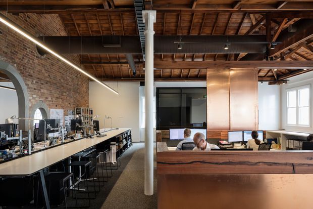 Interior view of a contemporary open-plan office in a converted industrial building. The workspace features exposed red-brick walls, exposed timber roof rafters and steel beams, a long communal workbench with stools, individual desks, computers, a large copper-toned partition or service volume, and abundant natural light from tall windows.