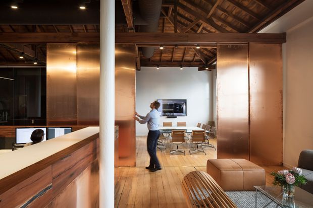 Warmly lit open-plan industrial-style interior combining a reception or work counter in the foreground with a meeting and lounge area beyond. The space features exposed timber roof framing, polished timber flooring, copper-toned metal wall panels, dark ceiling services, a wall-mounted screen, modern meeting chairs, a low lounge table, and leather seating.