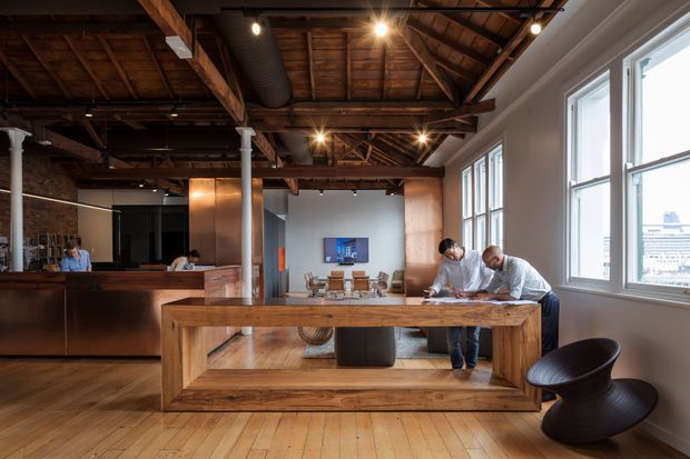 Open-plan loft-style interior with a large timber-framed communal table or workspace in the foreground, exposed dark timber roof structure and ductwork overhead, polished concrete and timber elements, a kitchen or bar area at left, and tall industrial-style windows along the right wall. Several people are working around the table, with a lounge or dining area visible beyond.