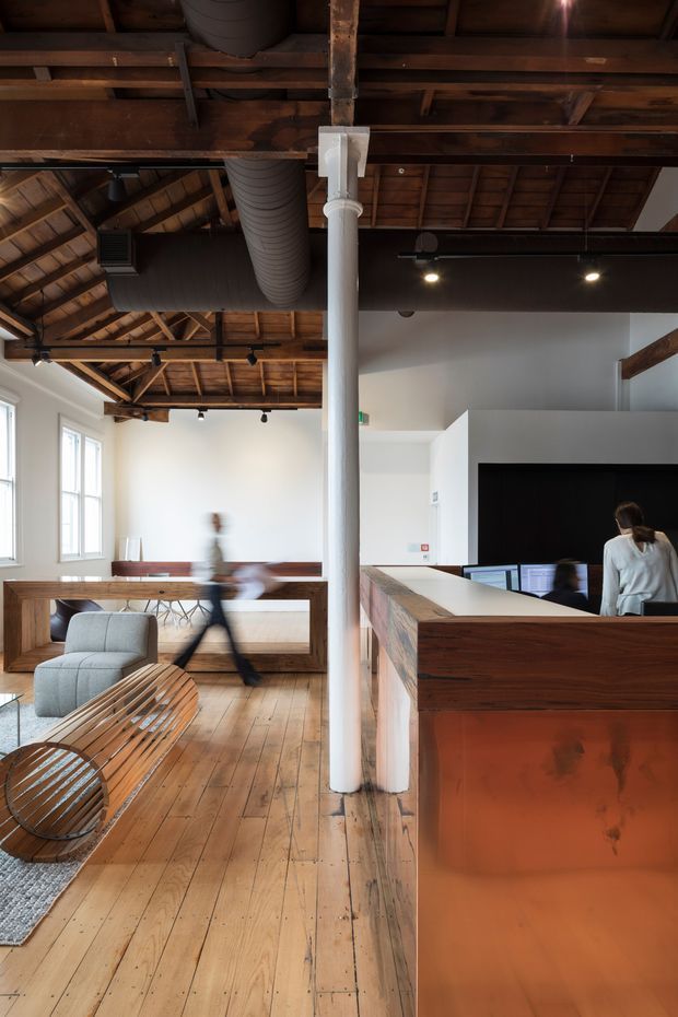 Interior view of a contemporary commercial reception and lounge space in a converted industrial or heritage building. The room features exposed timber roof structure and rafters, polished? worn timber plank flooring, large white-painted walls, exposed dark mechanical ductwork, track lighting, a central white column, timber built-in seating and counter joinery, and a copper-toned reception desk. Blurred people are present, with lounge seating in the foreground and a large black display wall at reception.