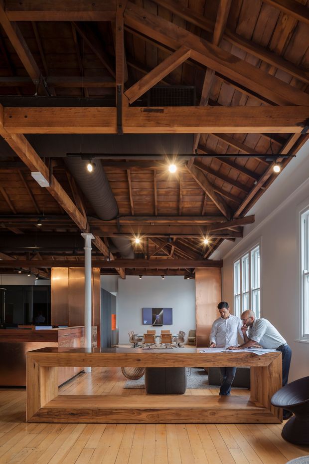 A warm, open-plan commercial interior with exposed timber roof trusses, rafters and beams, polished-looking timber flooring, and large multi-pane windows. A substantial timber reception or work counter occupies the foreground, with two people reviewing plans. The space includes exposed ductwork, track and pendant-style lighting, copper-toned wall panels, a dining or meeting area, and a contemporary office fit-out within an industrial warehouse-like shell.