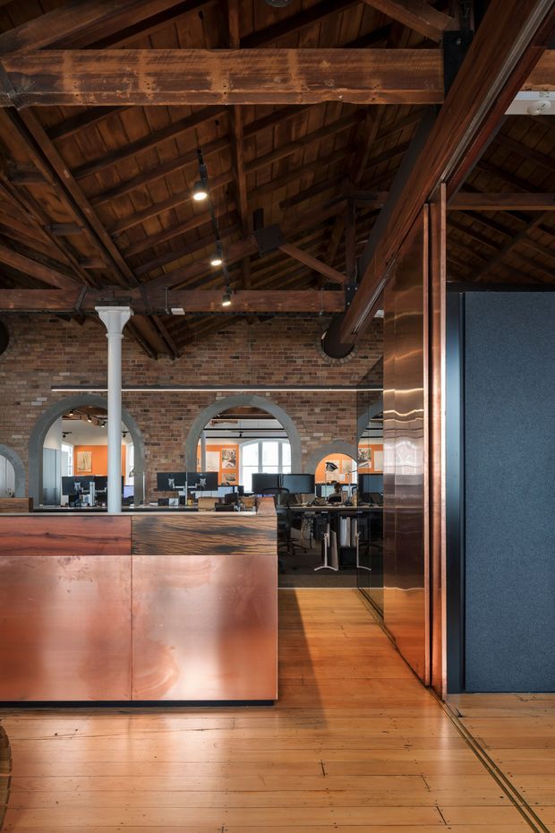 A warm, open-plan commercial office interior in a converted brick building. The space features exposed dark timber roof trusses and rafters, weathered brick walls with large arched openings, timber flooring, copper-toned wall and reception counter cladding, and contemporary workstations visible beyond the reception area. A dark blue acoustic or partition wall forms the right side of the image.