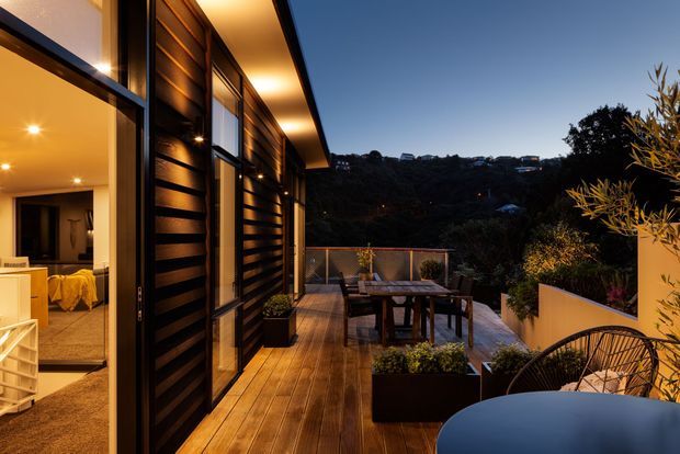A contemporary timber-clad home viewed from a timber deck at dusk. Warm interior lighting is visible through large glazed openings, while the deck features outdoor dining furniture, planters, and a lounge chair overlooking a wooded hillside.