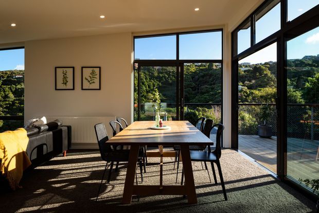 A bright contemporary dining and living area with a timber dining table surrounded by black chairs, a partially visible dark sofa, framed botanical artwork, and extensive dark-framed glazing. The windows open onto a deck or balcony overlooking dense greenery, with strong afternoon sunlight casting shadows across the carpeted interior.