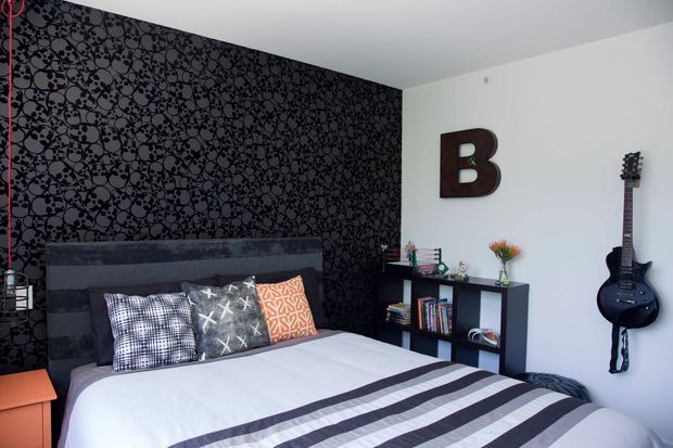 A stylish contemporary bedroom with a large bed, dark upholstered headboard, patterned monochrome feature wallpaper, and striped bedding. A black open shelving unit displays books and decor, while a wall-mounted guitar and oversized letter B add personality. The room combines dark graphic accents with a bright white wall and ceiling.
