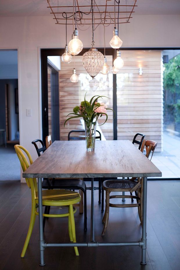 A dining room centered on a large rectangular timber dining table with exposed metal pipe-style legs, surrounded by mismatched chairs in yellow, black, and natural timber tones. A cluster of pendant lights, including a prominent crystal-like chandelier, hangs from an exposed ceiling grid. Full-height glazing and timber cladding are visible beyond the table, creating a bright connection to the exterior.