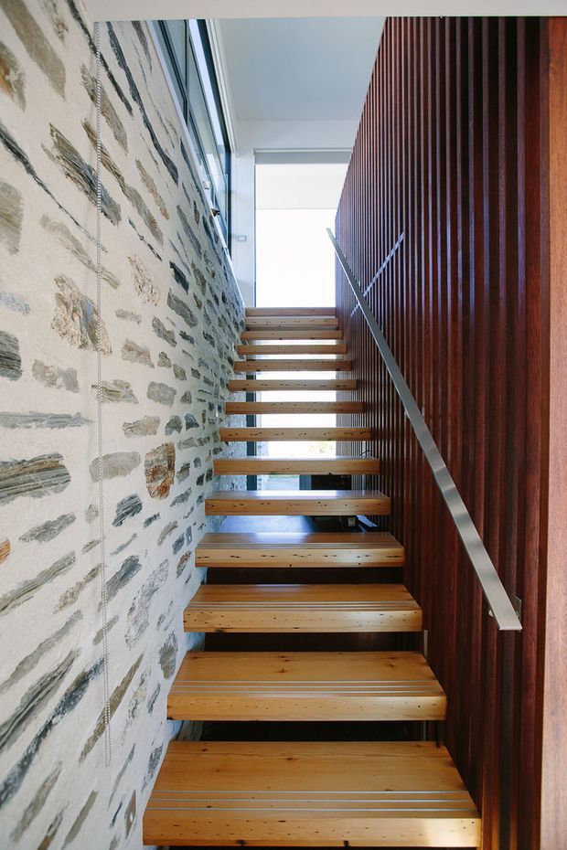 A narrow, modern stairway rises between a rough exposed stone wall and a dark vertical timber slat wall. Open-riser timber treads are paired with a slim metal handrail, with bright daylight entering from the upper landing and glazing beyond.