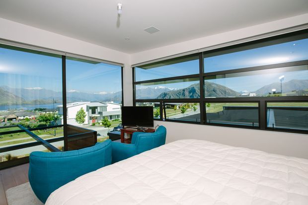 Bright contemporary bedroom with a large bed in the foreground, vivid turquoise upholstered seating, and extensive black-framed corner glazing. The windows provide panoramic views across a lake and mountainous landscape, while a television and small timber table occupy the seating area. The room has a simple white ceiling and walls with dark timber-toned flooring.