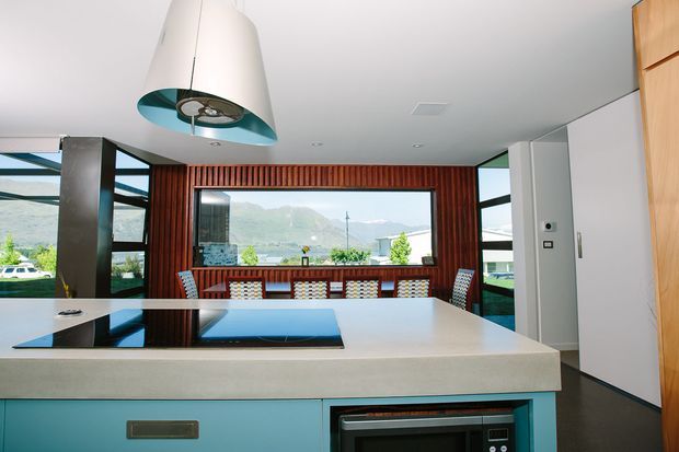 Contemporary kitchen viewed from the island, featuring a large pale benchtop with integrated black cooktop, turquoise-blue island cabinetry, light timber cabinetry, a sculptural pendant light, and a dining nook framed by dark red timber battens. Expansive glazing provides views toward a mountainous landscape.