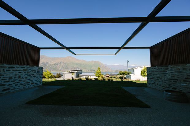A covered outdoor courtyard or terrace framed by dark steel beams, with low stone walls and timber privacy screens on either side. The open roof structure creates a strong view toward a sunlit mountain landscape, with landscaped lawn and paved ground in the foreground.