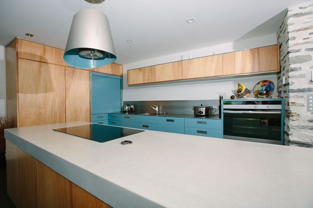 A contemporary kitchen featuring extensive light timber cabinetry, turquoise-blue lower cabinets and splashback, a large pale island benchtop with integrated cooktop, stainless-steel appliances, and a large sculptural pendant rangehood. A textured exposed stone wall forms a strong feature at the right.