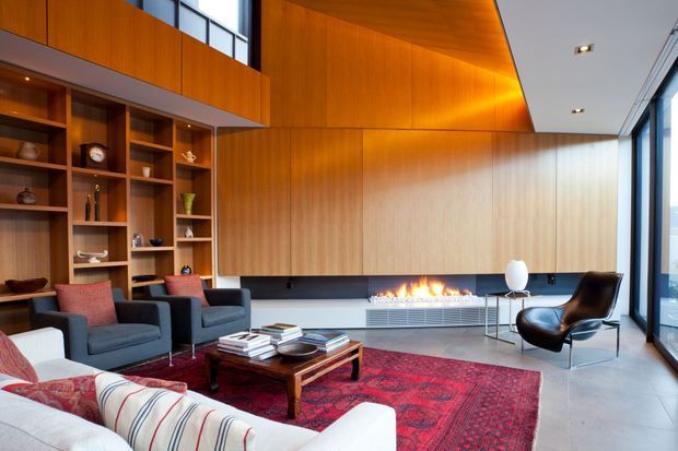 A spacious contemporary living room with extensive warm timber wall and ceiling panelling, built-in open shelving, a long linear fireplace, and floor-to-ceiling glazing. The room contains dark lounge chairs, a white sofa, a low timber coffee table, and a large red patterned rug, creating a refined yet warm atmosphere.