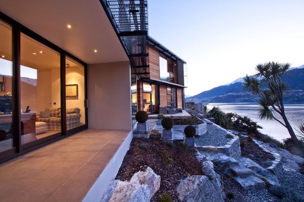 Contemporary multi-level residence on a rocky lakeside site, viewed from a covered terrace. The home features extensive dark-framed glazing, warm timber cladding, projecting upper volumes, and a distinctive overhead metal screen or trellis. Landscaped stone terraces and sculpted planting lead toward the water and mountainous backdrop. A warmly lit living room is visible through the glazing.