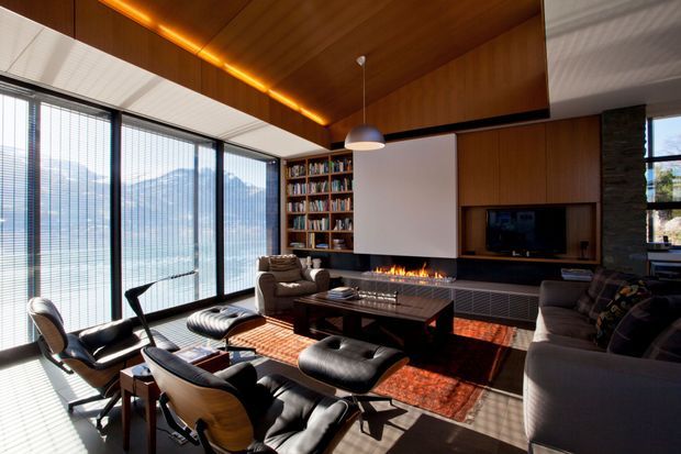 A sophisticated mid-century-inspired living room with expansive floor-to-ceiling glazing, mountain views, warm timber wall and ceiling panelling, a built-in bookcase, fireplace, television joinery, leather lounge chairs, a large sectional sofa, and a central coffee table. The room combines rich timber, dark furnishings, and a patterned rug with abundant natural light.