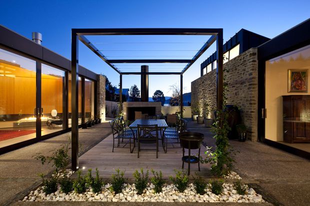 A contemporary courtyard viewed from outdoors, centred on a timber deck dining area beneath a dark steel pergola with a transparent roof. Flanking wings feature extensive glazing, dark metal framing, warm interior lighting, and textured stone walls. Gravel, white decorative stones, and low planting border the courtyard, with a built-in outdoor fireplace or hearth at the rear.
