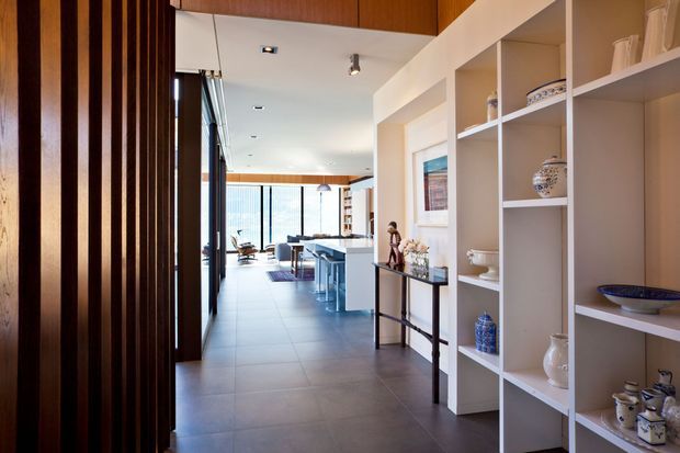 A bright contemporary open-plan residential interior viewed along a hallway toward a kitchen and living area. The space features dark vertical timber slats at the entry, large-format grey tiled flooring, white built-in display shelving, timber overhead cabinetry and ceiling accents, and expansive glazing at the far end. Decorative ceramics occupy the shelving.