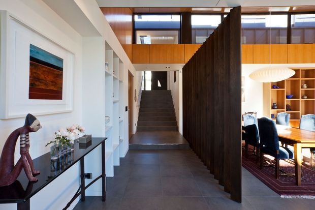 A bright contemporary residential entry hall with a long dark tiled floor, white built-in shelving and a slim console on the left, and a dramatic dark timber screen dividing the circulation space from a dining room. A staircase rises toward a mezzanine or upper-level landing, with warm timber wall panelling, clerestory glazing, and a large sculptural pendant visible over the dining table.