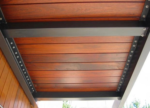 Close-up upward view of a timber-lined exterior overhead structure, likely a covered patio or porch roof. Warm reddish-brown timber boards are arranged between dark exposed metal or painted structural beams with visible bolted fixings. A timber-clad wall and bright outdoor surroundings are visible at the edges.