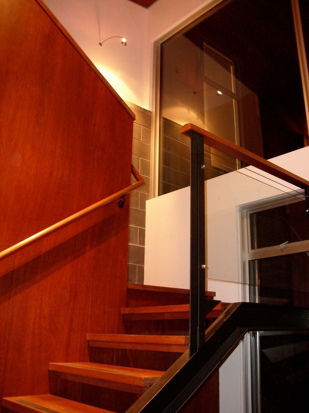 Interior view looking up a compact multi-level stairway with rich reddish timber stair treads, a tall timber-clad wall and timber handrail, black metal balustrade posts and rails, and white plasterboard walls. A large upper-level window or glazed opening and warm spot lighting create a dramatic, warmly lit circulation space.