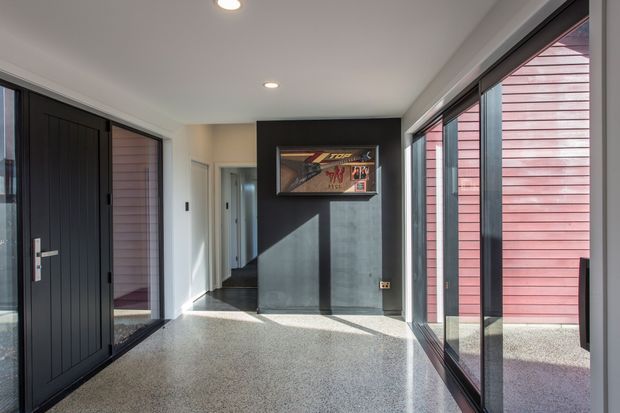 A bright contemporary entry hall with a polished speckled floor, dark vertical-panel front door, full-height black-framed glazing, and a dark feature wall displaying framed artwork. Pink-red horizontal exterior cladding is visible through the glazing, and recessed ceiling lights illuminate the space.