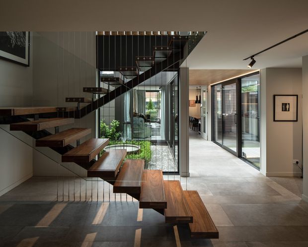 A refined contemporary interior centred on a floating timber staircase with dark metal stringers and slender vertical balustrade wires. The staircase rises beside a glazed internal courtyard or garden, while a long hallway with expansive black-framed glazing extends to the right. Large-format pale floor tiles, warm timber flooring, neutral walls, integrated track lighting, and abundant natural light create a sophisticated, open circulation space.