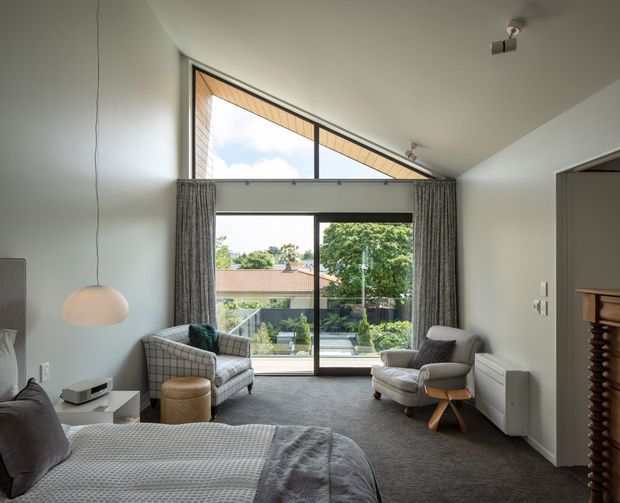 A bright, spacious bedroom with a high sloping ceiling and dramatic triangular clerestory glazing above full-height sliding glass doors. The room contains a bed, two upholstered armchairs, a small ottoman, side tables, a wall-mounted television or console, curtains, and carpet flooring. The glazing overlooks a leafy residential garden and neighbouring homes. Neutral grey and white finishes create a calm, contemporary atmosphere.