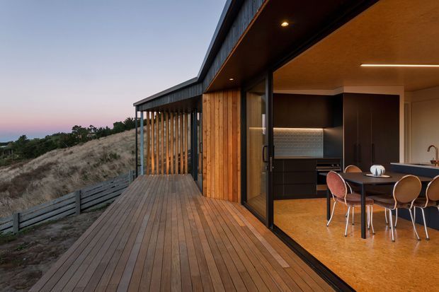 A contemporary timber-clad dwelling with a broad timber deck and open glazed corner overlooking a natural coastal or rural landscape at dusk. The interior visible through the fully opened glazing includes a compact kitchen and dining area, with dark cabinetry, timber ceiling, warm flooring, and pendant-free linear lighting. Vertical timber screening and a low-slung roof create a strong indoor-outdoor connection.