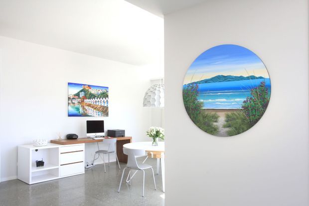Bright minimalist home office and dining nook with white walls, polished grey concrete flooring, a compact timber-and-white desk with computer, white shelving, a small round dining table, and white chairs. A large circular artwork or window-like wall feature depicts a vivid coastal beach and mountain scene, while a sculptural pendant light hangs above the workspace. The room has a clean, airy, contemporary feel.