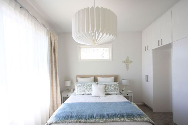 Bright, softly styled bedroom with a centered double bed, pale blue throw, patterned cushions, white bedside tables and lamps, full-height sheer curtains with beige drapes, and extensive built-in white wardrobes. A sculptural pleated pendant light and small decorative wall cross add visual interest. The room has a calm, airy, refined character with light neutral finishes and carpeted flooring.