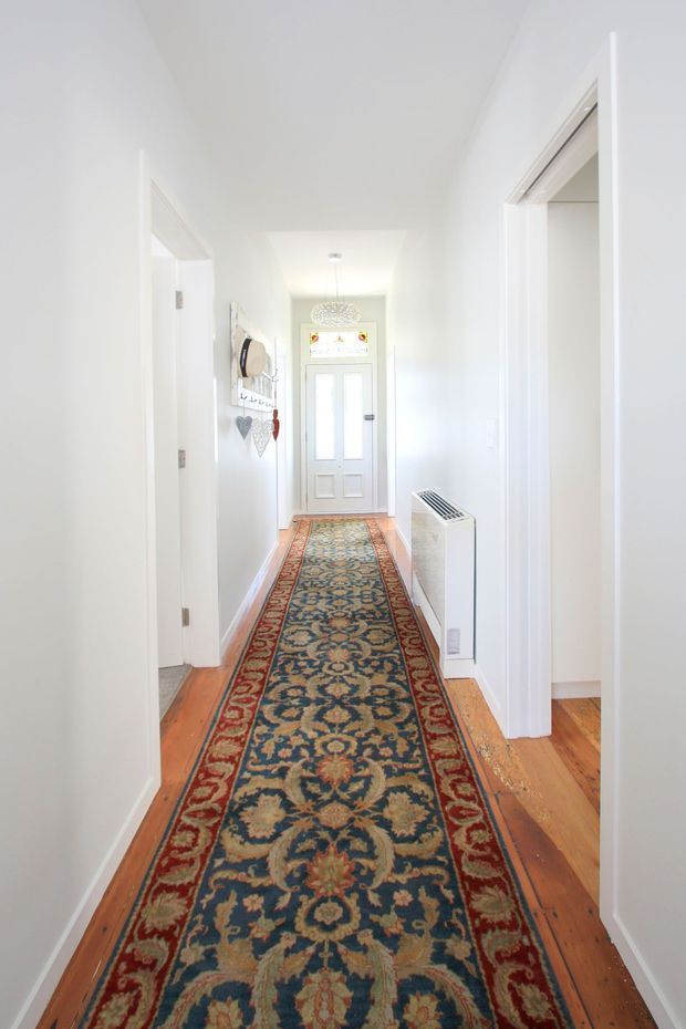Bright, narrow residential hallway with white walls and trim, warm timber floorboards, and a long traditional patterned runner rug. Several doorways open off the corridor, leading to other rooms, and a white entry door with decorative glazing is visible at the far end. A compact white wall heater and small decorative wall hanging are also present.
