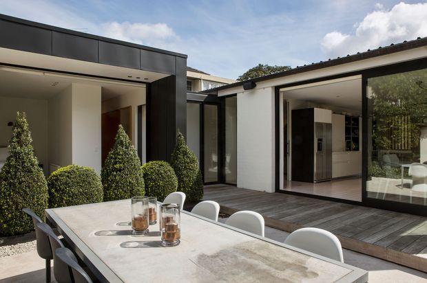 Contemporary single-storey residence with a sheltered courtyard terrace and outdoor dining table in the foreground. Dark charcoal cladding and trim contrast with white rendered walls, while extensive sliding glass doors open to a bright kitchen and living area. Sculpted evergreen planting borders the timber deck, creating a refined indoor-outdoor connection.