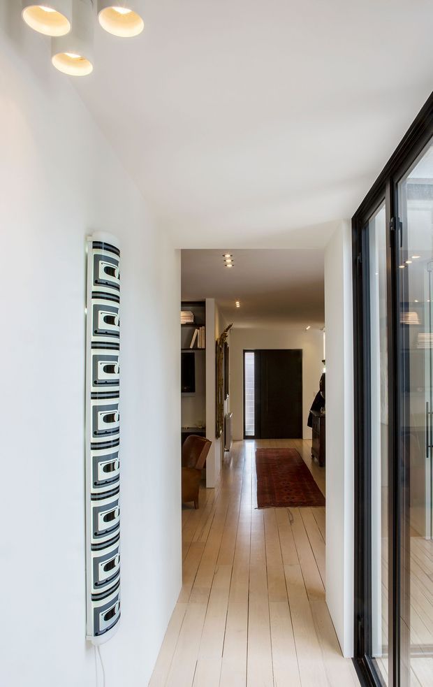 A bright, narrow residential hallway or entry passage with pale timber plank flooring, smooth white walls and ceiling, recessed and oval ceiling lights, and a dark-framed glazed opening along the right side. A patterned vertical wall-mounted decorative feature is installed on the left, while the corridor leads toward a dark entrance door and a timber-floored living area with a runner rug and glimpses of furniture.