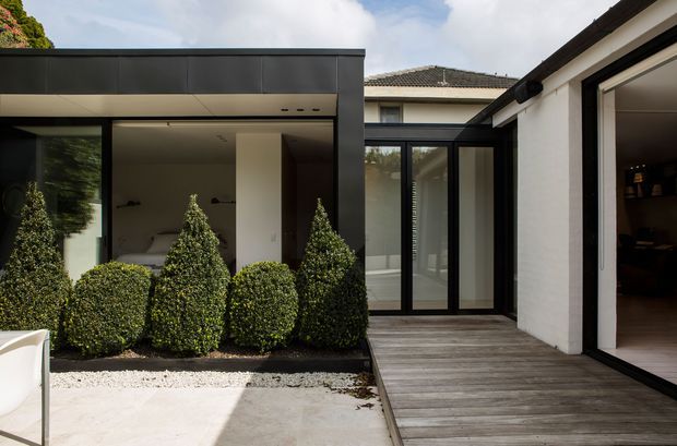 A contemporary courtyard renovation viewed from outdoors, with a dark flat-roofed pavilion on the left, black-framed floor-to-ceiling glazing and doors, a weathered timber deck, pale concrete paving, and neatly clipped evergreen shrubs in a gravel garden bed. The courtyard connects to a bright interior space and a white-rendered building with a tiled roof visible beyond.