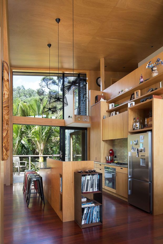 A warm, compact open-plan kitchen with extensive honey-toned timber cabinetry, shelving, wall and ceiling lining, and dark polished timber flooring. The kitchen includes a peninsula with bar stools, integrated shelving, a stainless-steel refrigerator, tiled splashback, oven and benchtop appliances. Full-height glazing opens to lush tropical planting, while black-framed windows and sculptural pendant lights add contrast. A double-height or loft-like volume is suggested above the kitchen, with a small upper opening and visible stair or landing area.