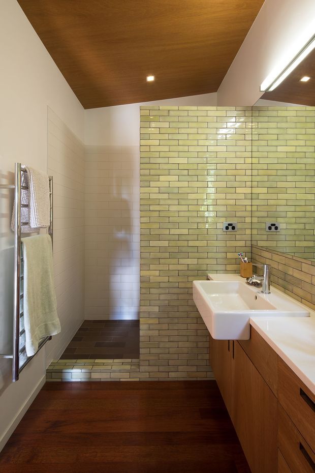 A compact contemporary bathroom with a long timber vanity and white countertop, twin white wall-mounted basins, chrome tapware, and a large mirrored wall. Glossy pale green brick-pattern tiles form the main feature wall and continue through the shower area, while the adjacent walls are tiled in soft off-white. Dark timber flooring and a warm timber ceiling create a cohesive, earthy palette. A chrome heated towel rail is visible beside the shower.