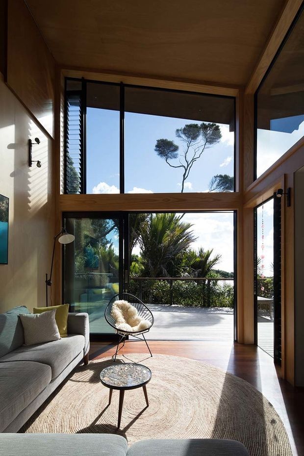 A sunlit modern living room with a double-height timber-lined ceiling and expansive black-framed glazing opening to a balcony or terrace. The room features a light grey sofa, sculptural lounge chair, round coffee table, and large circular natural-fibre rug. Lush planting and mature trees are visible beyond the glass, creating a strong indoor-outdoor connection.