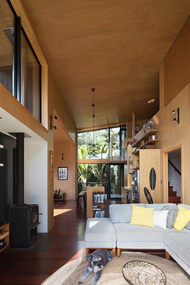 A warm, double-height living room with extensive timber wall and ceiling lining, dark timber flooring, and floor-to-ceiling glazing overlooking lush vegetation. The room includes a large pale sectional sofa, black wood-burning stove, suspended pendant lights, open built-in shelving, a timber door with decorative oval inlay, and a staircase. A mezzanine-like upper level and tall glazed opening emphasize the vertical volume.