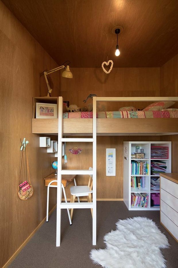 Compact children's bedroom with a built-in loft bed and white ladder, enclosed in warm timber wall and ceiling panelling. The lower level includes a small desk and chair, open shelving filled with books and craft materials, and white drawer cabinetry. A colourful patchwork quilt, children's artwork, hanging accessories, pendant bulb, desk lamp, and white fluffy rug create a playful, cosy atmosphere.